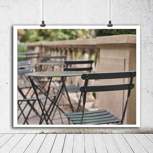 May include: A black metal bistro table with four black metal chairs on a patio. The table and chairs are set up in a row, with the chairs facing forward. The background is blurred and shows a stone wall and green foliage.