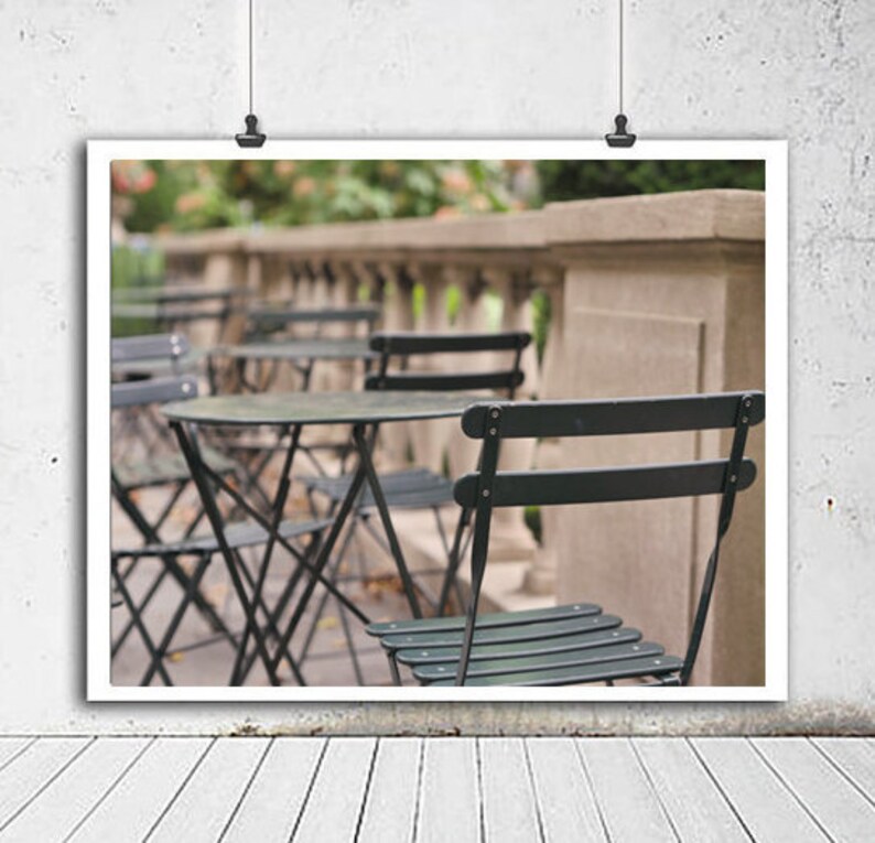 May include: A black metal bistro table with four black metal chairs on a patio. The table and chairs are set up in a row, with the chairs facing forward. The background is blurred and shows a stone wall and green foliage.