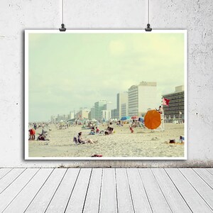 May include: A vintage photograph of a beach scene with people relaxing on the sand. The photo features a large orange beach umbrella, a lifeguard stand, and a row of buildings in the background. The sky is a light blue with a few clouds.