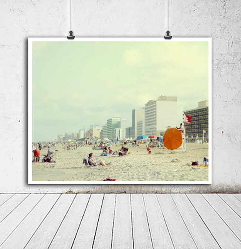 May include: A vintage photograph of a beach scene with people relaxing on the sand. The photo features a large orange beach umbrella, a lifeguard stand, and a row of buildings in the background. The sky is a light blue with a few clouds.