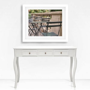 May include: A white console table with four drawers and curved legs. The table is in front of a white wall with a framed photograph of a row of black metal chairs and tables on a patio.