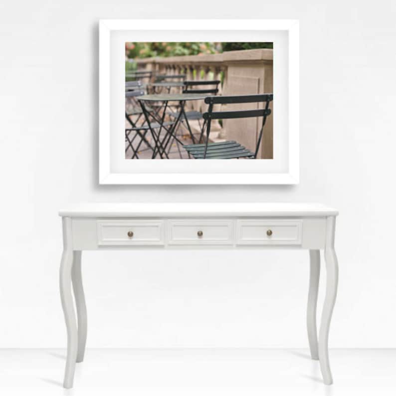 May include: A white console table with four drawers and curved legs. The table is in front of a white wall with a framed photograph of a row of black metal chairs and tables on a patio.