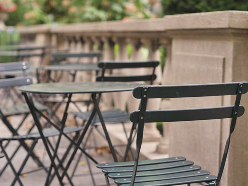 May include: A set of black metal bistro tables and chairs on a patio. The tables are round and have a black metal frame. The chairs are also black metal and have a slatted seat.