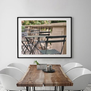 May include: A black and white photograph of a Parisian cafe with black metal tables and chairs. The cafe is located on a balcony with a stone railing. The photo is framed in a black frame.