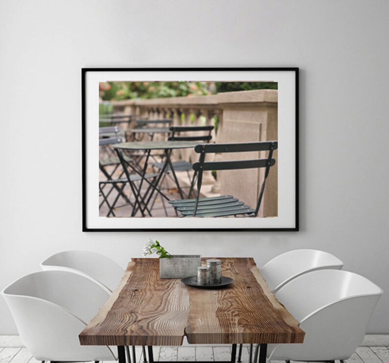 May include: A black and white photograph of a Parisian cafe with black metal tables and chairs. The cafe is located on a balcony with a stone railing. The photo is framed in a black frame.