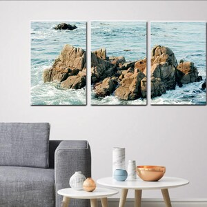 Może przedstawiać: Trzyczęściowy wydruk na pł&oacute;tnie przedstawiający krajobraz morski z dużymi skałami i rozbijającymi się falami. Skały są jasnobrązowe, a woda jest ciemnoniebieska.