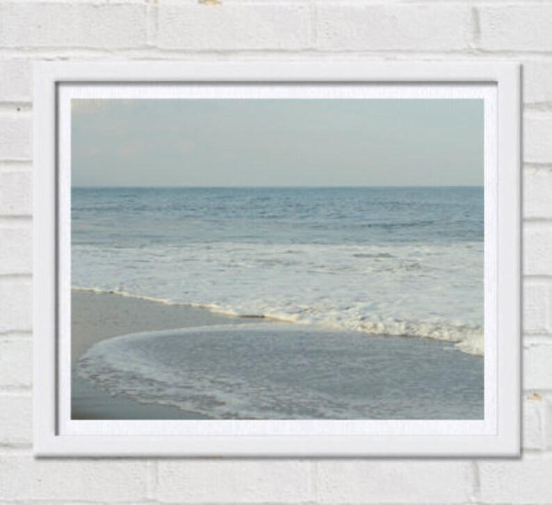 May include: A framed photograph of a beach scene with white foamy waves crashing on the shore. The water is a light blue color and the sky is a pale blue.