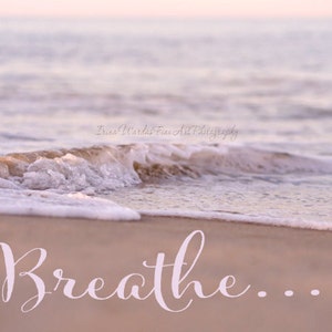 May include: A close-up of a foamy wave crashing on a sandy beach. The water is a soft pink and the sand is a warm beige. The word "Breathe..." is written in white cursive on the sand.