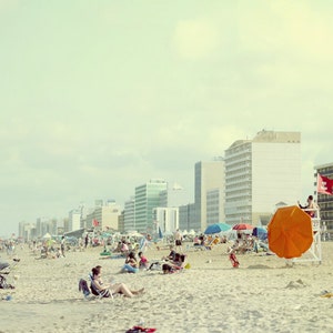 May include: A crowded beach with a lifeguard stand and an orange umbrella. There are several buildings in the background, including a tall white building with a red flag.