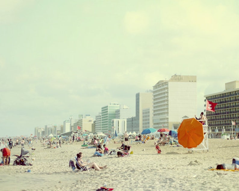 May include: A crowded beach with a lifeguard stand and an orange umbrella. There are several buildings in the background, including a tall white building with a red flag.