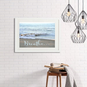 May include: A white framed print with a photograph of a wave crashing on a sandy beach. The text "Breathe..." is written in blue cursive on the sand.
