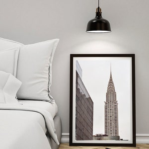 May include: A black framed print of a skyscraper in a city. The building is tall and narrow with a pointed top. The print is hanging on a white wall above a white bed with white pillows.