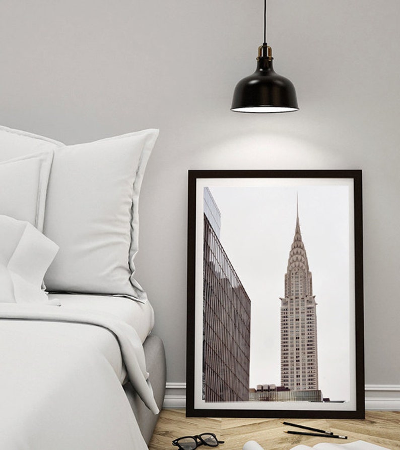May include: A black framed print of a skyscraper in a city. The building is tall and narrow with a pointed top. The print is hanging on a white wall above a white bed with white pillows.