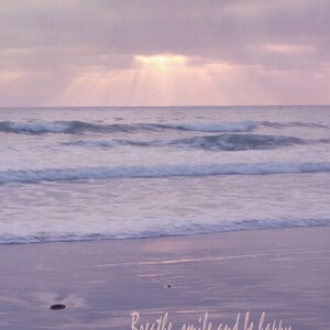 Pode incluir: Um suave p&ocirc;r do sol rosa e roxo sobre um oceano calmo com ondas que rolam suavemente para uma praia de areia. O texto "Breathe, smile and be happy..." est&aacute; escrito na areia.