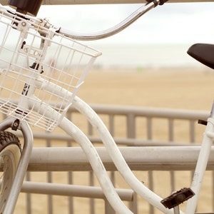 Puede incluir: Una bicicleta blanca con una cesta y un asiento negro. La bicicleta est&aacute; aparcada en un paseo mar&iacute;timo de madera.
