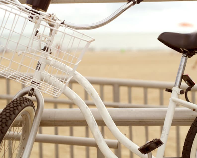 Puede incluir: Una bicicleta blanca con una cesta y un asiento negro. La bicicleta est&aacute; aparcada en un paseo mar&iacute;timo de madera.