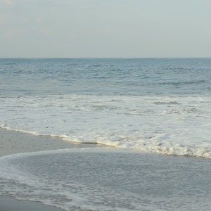 May include: A close-up of a sandy beach with foamy white waves crashing on the shore. The water is a light blue color and the sky is a pale gray.