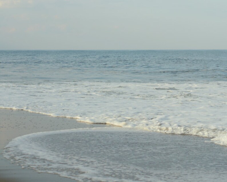 May include: A close-up of a sandy beach with foamy white waves crashing on the shore. The water is a light blue color and the sky is a pale gray.