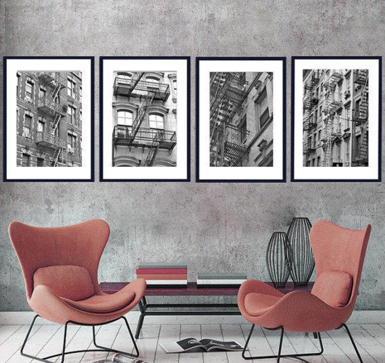 May include: Four black framed photographs of black and white images of buildings with fire escapes. The photographs are hanging on a gray wall. Two red armchairs are in the foreground.