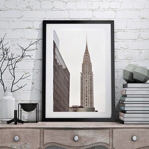 May include: A black framed print of a skyscraper in a city. The print is on a wooden shelf with a vase, a bowl, and a stack of books.