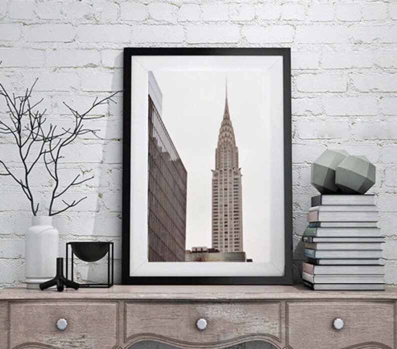 May include: A black framed print of a skyscraper in a city. The print is on a wooden shelf with a vase, a bowl, and a stack of books.