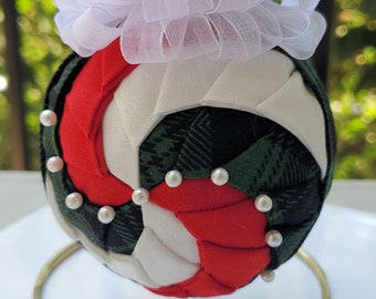 May include: A handcrafted Christmas ornament with a red, white, and green plaid design. The ornament is adorned with small pearls and topped with a red and white bow. It hangs from a natural twine string.