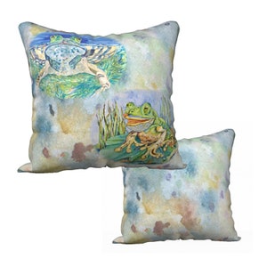 Könnte beinhalten: Zwei dekorative Kissen mit Aquarellhintergrund und Froschmotiv. Ein Kissen zeigt zwei Frösche, die auf einem Seerosenblatt sitzen, während das andere Kissen einen einzelnen Frosch zeigt, der auf einem Seerosenblatt sitzt.
