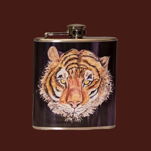 Puede incluir: Una petaca de plata con un diseño de cara de tigre. El tigre tiene rayas naranjas y negras y un hocico blanco. La petaca tiene una tapa de plata.