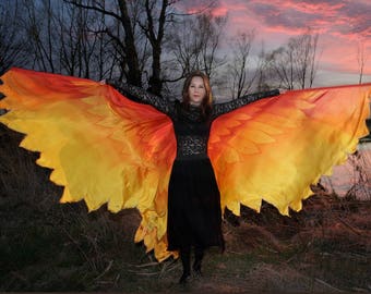 Può includere: Una donna con un vestito nero tiene un grande tessuto fluido con un gradiente di arancione e giallo. Il tessuto ha un motivo a forma di foglia ed è tenuto davanti a lei. Lo sfondo è un'immagine sfocata di alberi e un cielo al tramonto.