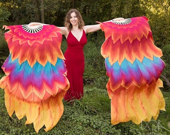 May include: A woman in a red dress holds two large, colorful fabric fans. The fans are decorated with a pattern of orange, yellow, red, pink, and blue feathers. The woman is standing in a grassy area with trees in the background.