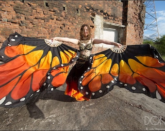 May include: A woman wearing a black costume with gold accents is holding two butterfly-shaped fans. The fans are orange, yellow, and black with white spots. The woman is standing in front of a brick wall.