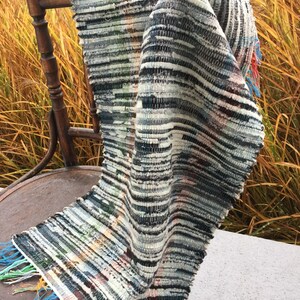 May include: A handwoven textile rug with a striped pattern in shades of gray, white, and blue, draped over a wooden chair. The rug has fringed edges with colorful threads. The chair is made of dark wood.