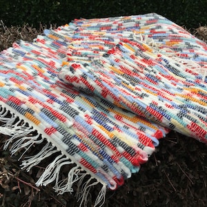 May include: A colorful, woven rug with a rectangular shape. The rug features a mix of red, white, blue, yellow, and gray threads, creating a vibrant pattern. The edges are finished with a white fringe. This handmade rug is suitable for home decor.