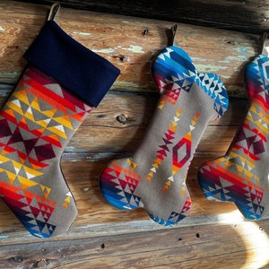 Op de afbeelding: Drie kerstsokken met een geometrisch patroon uit het zuidwesten in tinten rood, oranje, geel en blauw. De sokken hebben een grijze basis en een marineblauwe manchet. Elke sok hangt aan een leren lus.