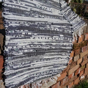 May include: A rectangular, handwoven rug in shades of white, grey, and navy blue, with a fringed edge. The rug is displayed on a red brick wall. The woven texture of the rug is clearly visible, highlighting the pattern.