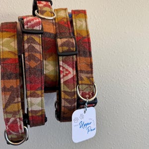 May include: A collection of dog collars with a colorful geometric pattern in shades of red, orange, yellow, and green. Each collar has a black buckle and a silver metal D-ring. A white tag with the text "The Upper Paw" hangs from one collar.