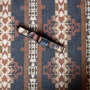 May include: A dog collar with a geometric pattern in shades of brown, beige, and grey, featuring a black plastic buckle and a silver metal ring. The collar is set against a matching patterned background.