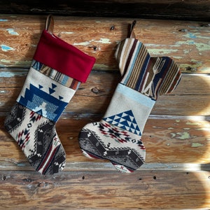 May include: Two Christmas stockings with a Southwestern design. One is a traditional shape with a burgundy cuff, the other is bone-shaped. Both have leather hanging loops and are made of patterned fabric in shades of beige, blue, and brown.
