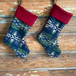 May include: Two Christmas stockings with a southwestern pattern in blue, green, and white. The top of each stocking is a solid burgundy color. The stockings are hanging on a wooden surface.