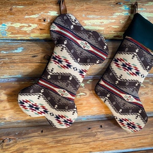 May include: Two Christmas stockings with a Southwestern pattern in brown, cream, red, and teal. One stocking is shaped like a bone, the other is a traditional stocking with a green cuff. Both are hanging on a rustic wooden background.