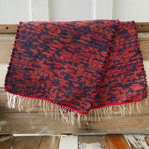 May include: A woven textile featuring a geometric pattern in red and navy blue. The edges are finished with a red border and white fringe. The textile is folded, showcasing its texture and design.