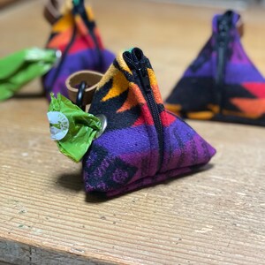 May include: Triangular dog waste bag holders with a colorful geometric pattern in purple, orange, and black. Each holder has a black zipper and a brown leather loop. A green bag of waste bags is attached to one holder.
