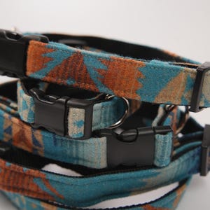 Puede incluir: Primer plano de varios collares para mascotas apilados. Los collares presentan un patrón geométrico turquesa y naranja con hebillas y herrajes de plástico negro. Los collares están hechos de un material suave y tejido.