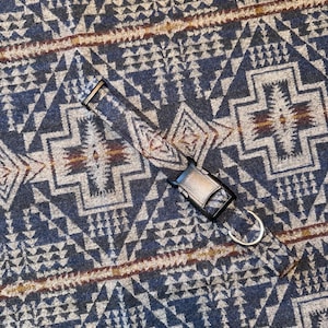 May include: A patterned dog collar with a black metal buckle and a silver metal D-ring. The collar features a geometric design in shades of blue, gray, and brown. The background is a matching fabric with the same pattern.