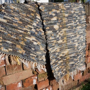 May include: Two rectangular woven rugs in shades of blue, beige, and cream, displayed against a brick wall. The rugs feature fringed edges and a textured weave, possibly handmade. The rugs are approximately 100 cm in length.