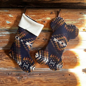 May include: Two Christmas stockings with a southwestern geometric pattern in navy blue, tan, and white. One stocking has a traditional shape, while the other is shaped like a dog bone. Both have a cream-colored cuff and leather hanging loop.