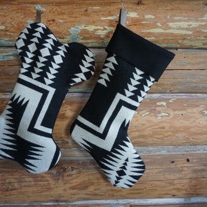 Op de afbeelding: Twee kerstsokken met een zwart-wit geometrisch patroon hangen tegen een rustieke houten achtergrond. De sokken hebben een omgeslagen zwarte manchet en een leren lus om op te hangen.