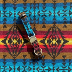 May include: A dog collar with a Southwestern-inspired pattern. The collar features a black, brown, and turquoise geometric design with a silver metal buckle and a D-ring. The background is a matching fabric with a similar pattern.