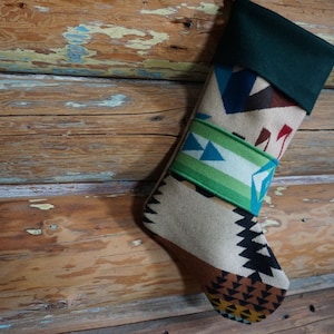 Può includere: Una calza di Natale con un design del sud-ovest. La calza presenta un polsino verde e un corpo a fantasia con forme geometriche nei toni del marrone, beige, rosso e blu. Una fascia verde e bianca con triangoli blu adorna la calza.