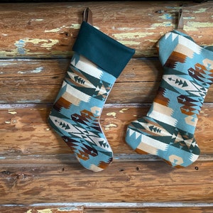 May include: Two Christmas stockings with a Southwestern pattern in shades of teal, brown, beige, and green. One stocking has a traditional shape, while the other is shaped like a dog bone. Both have a dark green cuff and a leather loop for hanging.